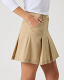 A model wearing a beige pleated mini skirt with side pockets and a white long-sleeve top, shown from the waist down against a light gray studio background.