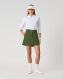 Daily Sports Woven Khakora Skort in green – women's A-line woven golf skort with structured waistband, silver logo button and side zip pocket, full length front view on model styled with white 3/4 sleeve polo, white visor and golf glove