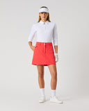 Daily Sports Woven Poppy Red Skort in poppy red – women's A-line woven golf skort with structured waistband, silver logo button and side zip pocket, full length front view on model styled with white 3/4 sleeve polo, white visor and golf glove