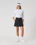 Daily Sports Woven Black Skort in black – women's A-line woven golf skort with structured waistband, silver double-button closure and side zip pocket, full length front view on model styled with white 3/4 sleeve polo, white visor and golf glove