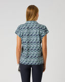 A woman viewed from behind wearing a short-sleeve collared top with a blue and white wavy geometric pattern, paired with dark pants, against a plain light gray studio background.