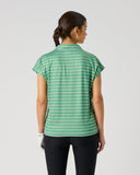 A woman viewed from the back wearing a green short-sleeve top with a white geometric pattern and black pants, standing against a light gray studio background.