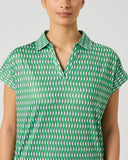 A woman wearing a green short-sleeve collared shirt with a repeating white geometric pattern, shown against a plain light gray studio background.