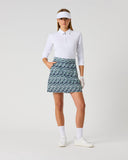 A woman modeling a white long-sleeve polo shirt and a blue and white patterned golf skirt, styled with a white visor, white gloves, white socks, and white golf shoes against a light gray studio background.