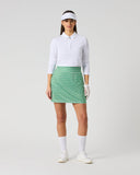 A woman modeling a white long-sleeve polo shirt and a green and white patterned mini skirt, paired with white socks, a white visor, and white shoes, against a light gray studio background.
