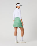 A woman modeling a green and white checkered mini skirt with a white long-sleeve top, white socks, white sneakers, and a white visor, photographed against a light gray studio background.