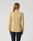 A woman viewed from the back wearing a beige long-sleeve mock-neck top and dark navy pants, standing against a plain light gray studio background.