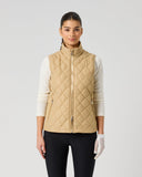 A woman modeling a quilted sleeveless vest in a light tan/beige color, worn over a cream long-sleeve shirt and black pants, against a plain light gray studio background.