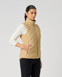 A woman modeling a beige quilted zip-up vest over a white long-sleeve top and black leggings, standing against a light gray studio background.