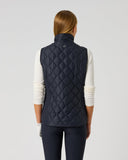 A woman viewed from the back wearing a quilted navy sleeveless puffer vest over a white long-sleeve top and dark pants, standing against a light gray studio background.