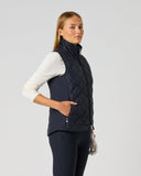 A woman modeling a navy blue quilted zip-up vest over a white long-sleeve top and dark leggings, standing against a light gray studio background.