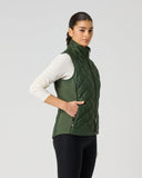 A woman modeling a dark green quilted sleeveless puffer vest over a white long-sleeve top and black leggings, standing against a light gray studio background.