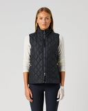 A woman modeling a black diamond-quilted zip-up vest over a white long-sleeve top and navy pants, standing against a light gray studio background.