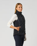 A woman modeling a black quilted sleeveless vest over a white long-sleeve top and dark leggings, standing against a light gray studio background.