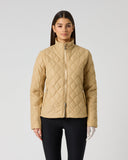 A woman modeling a quilted zip-up jacket in a light tan/beige color, paired with black pants, against a plain light gray studio background.