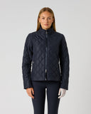 A woman stands against a light gray studio background wearing a dark navy quilted zip-front jacket with a stand collar, paired with dark leggings and white gloves.