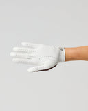 A white golf glove with perforated details and a small silver wrist emblem, worn on a person's hand against a solid light gray background.