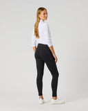 A woman standing in profile wearing black high-waisted leggings, a white long-sleeve top, and white sneakers against a light gray studio background.
