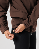 A person wearing a brown zip-front jacket and black leggings, shown from the chest down against a light gray studio background. The person is adjusting the hem of the jacket with both hands.