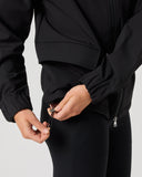 A close-up of a person wearing a black zip-front jacket and black leggings, adjusting the drawstring at the waist. The background is a clean, light gray studio backdrop.