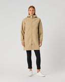 A woman modeling a beige hooded raincoat over black pants and white sneakers, standing against a light gray studio background.