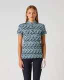 A woman modeling a short-sleeve collared golf shirt with a blue and white wavy geometric pattern, paired with dark pants and a white glove, against a light gray studio background.