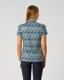 A woman viewed from the back wearing a short-sleeve collared shirt with a blue, white, and teal wavy geometric pattern, paired with dark navy pants, against a plain light gray studio background.