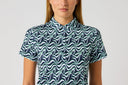 A woman wearing a short-sleeve collared button-up shirt with a wavy abstract pattern in navy blue, teal, and white, shown against a plain light gray background.