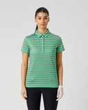 A woman modeling a short-sleeve polo shirt with a green and white geometric pattern, paired with black pants and a white glove, against a light gray studio background.