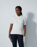 Woman wearing a white ribbed knit polo shirt and dark pants against a light gray background