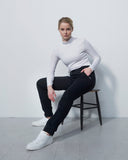 Person sitting on a stool wearing a white sweater and black pants against a plain background