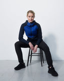 Person wearing a blue puffer vest sitting on a stool against a white wall.