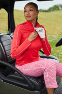A woman sits in a black golf cart on a grassy field, wearing a bright red long-sleeve zip-up athletic jacket with white piping and red-and-white gingham pants.