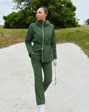 A woman stands on a sand bunker path outdoors wearing an olive green matching golf set consisting of a zip-up hooded jacket and matching pants. She is holding a golf club and wearing white golf shoes.
