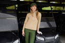 A woman stands in a golf cart wearing a beige long-sleeve zip-front athletic top, olive green pants, and an olive green cap. She is posed in front of the cart under a covered outdoor area.